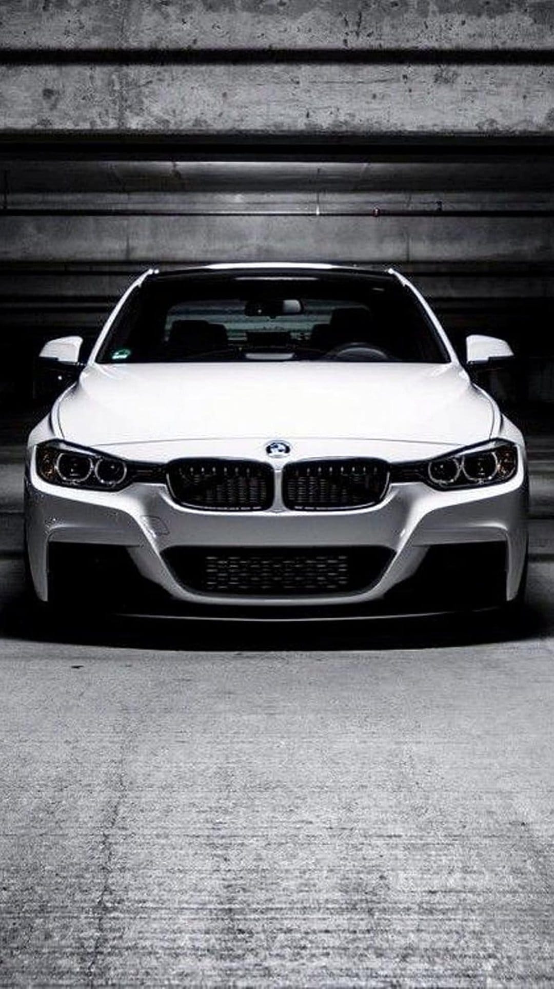 White BMW sedan with glowing headlights parked in a dark, industrial concrete garage with sharp shadows.