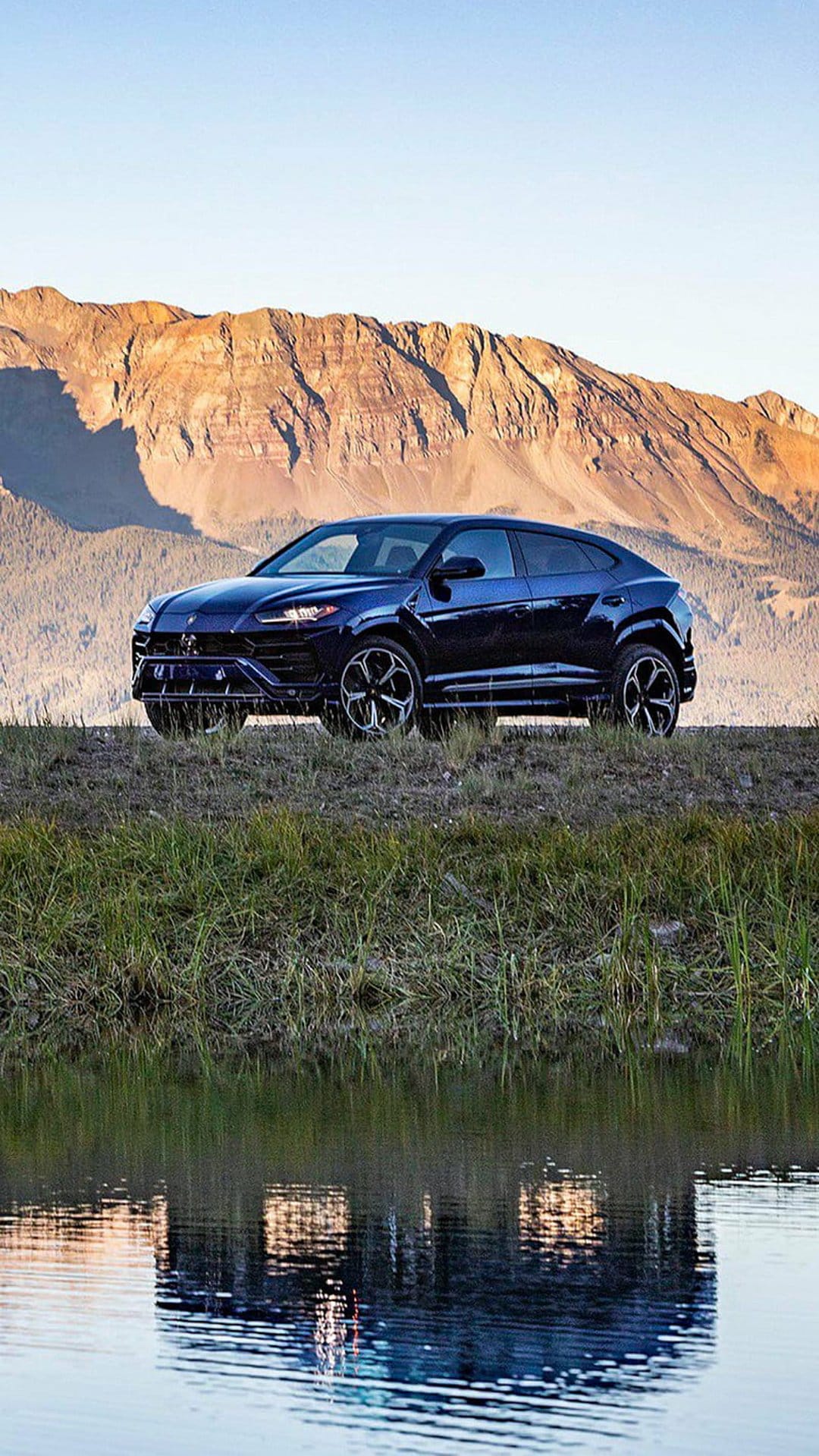 Dark blue Lamborghini Urus parked on a grassy ridge reflected in a calm mountain lake with snowy peaks.