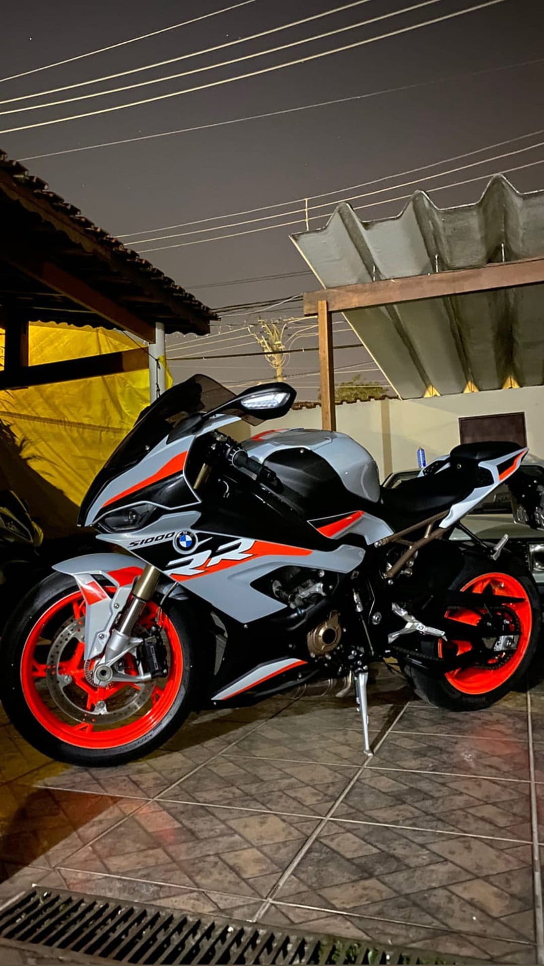 BMW S1000RR sportbike with orange wheels and grey racing livery parked on a tiled outdoor surface at night.