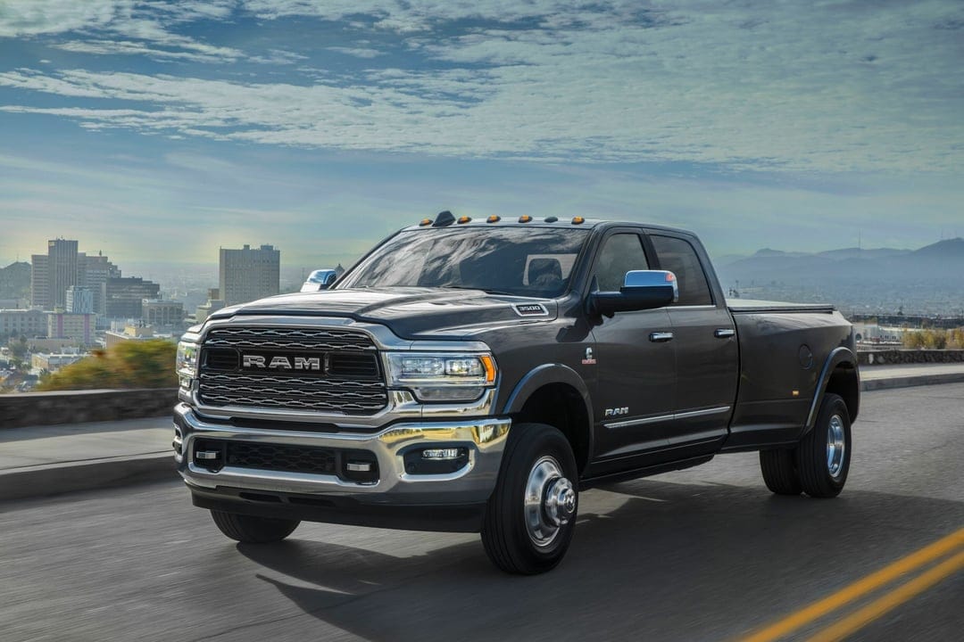 Dark gray Ram 3500 dually truck driving on a road with a city skyline and mountains under a blue sky.