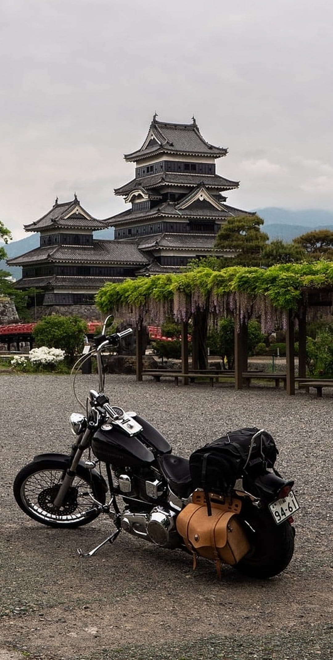 Black motorcycle parked on gravel beside a tiered Japanese castle under a canopy of purple wisteria blossoms.