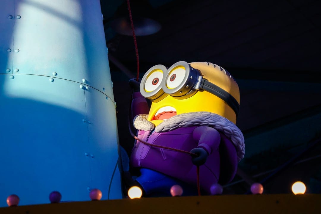 Minion wearing a purple winter jacket and goggles holds a red rope against a blue architectural background.