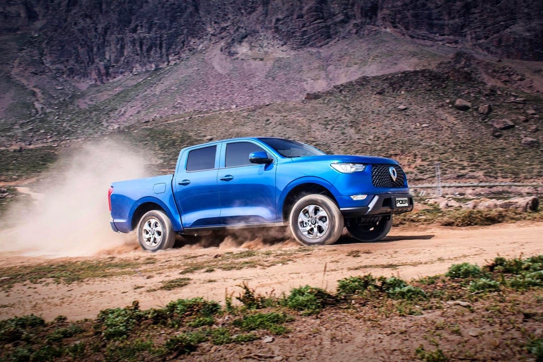 Blue pickup truck driving on a dirt road in front of rocky mountains, kicking up a thick cloud of dust.