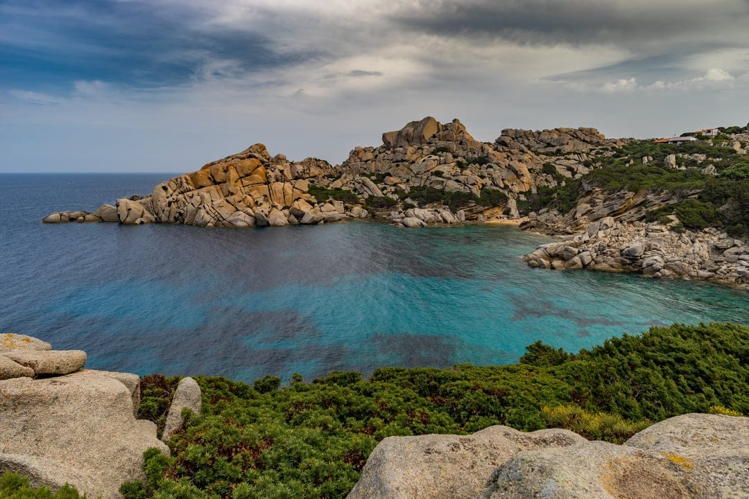 Sardinian granite cliffs overlook clear turquoise water and lush green hills under a bright mediterranean sky.