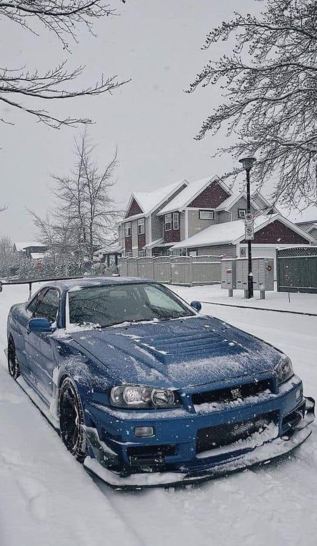 Blue Nissan Skyline R34 covered in white snow on a winter street with residential houses and bare trees.