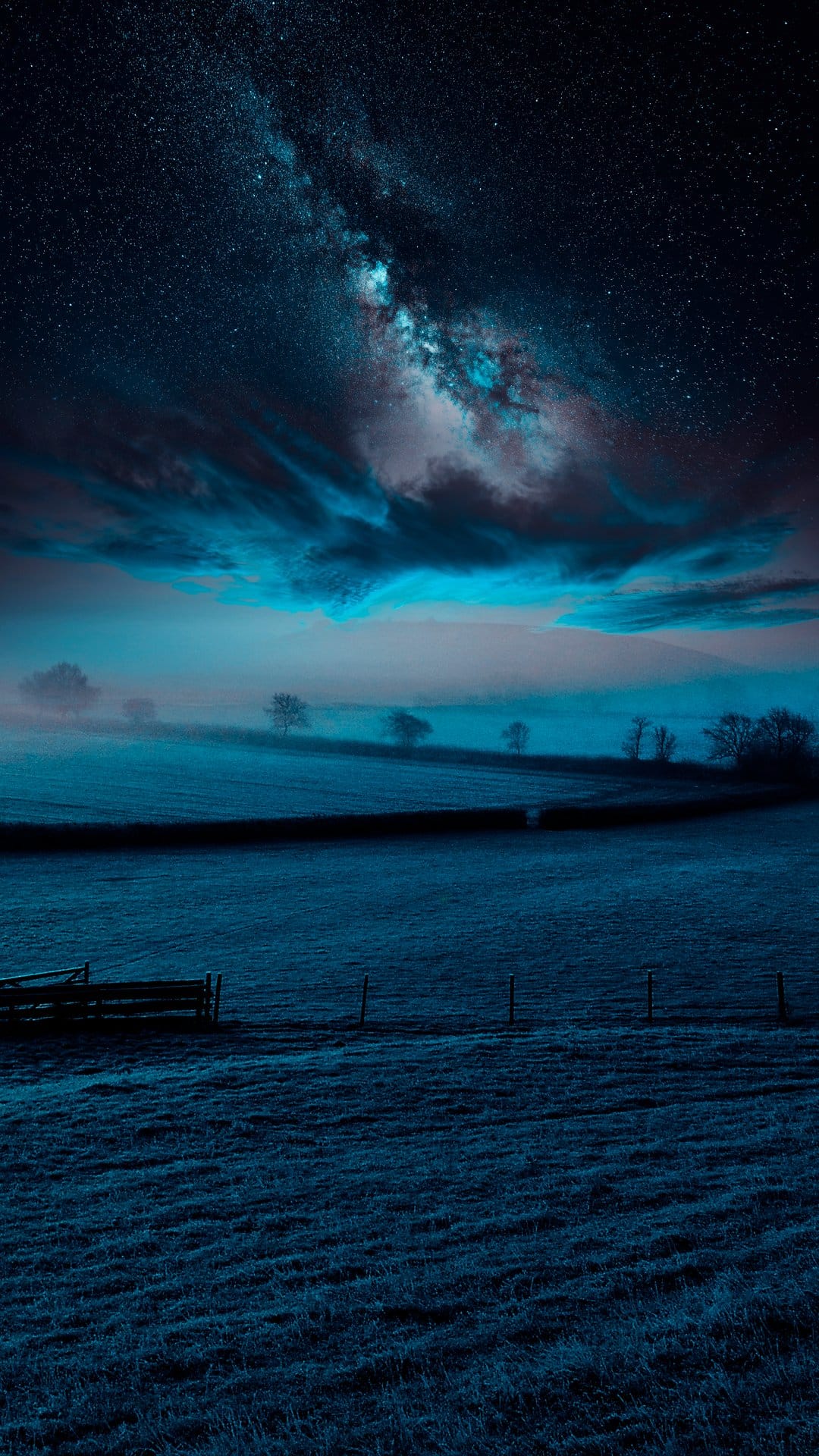 Milky Way galaxy core shines over a dense layer of ground fog and dark tree silhouettes in a blue night sky.