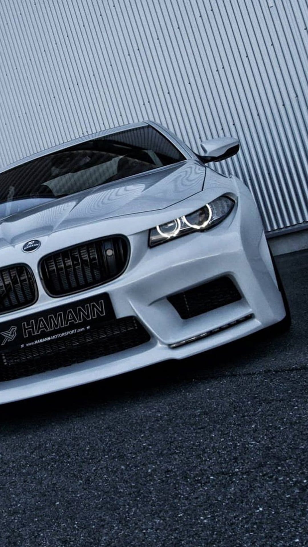 White BMW M5 with a Hamann front bumper and black grille parked against a grey corrugated metal wall on asphalt.