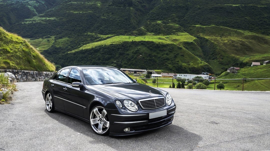 Dark luxury sedan parked on a winding road amidst sunlit green mountain peaks and a misty alpine valley.