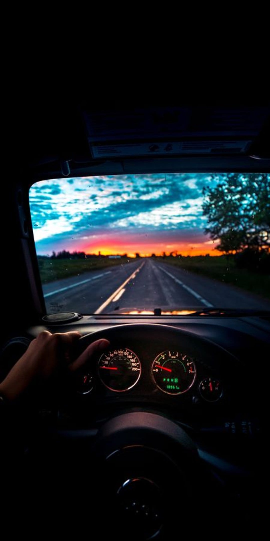 Hand on a steering wheel inside a car cabin facing a long road under a vibrant orange and blue sunset sky.