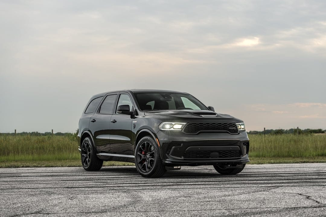 Black Dodge Durango SRT Hellcat with glowing headlights parked on a gray asphalt track under a cloudy sky.