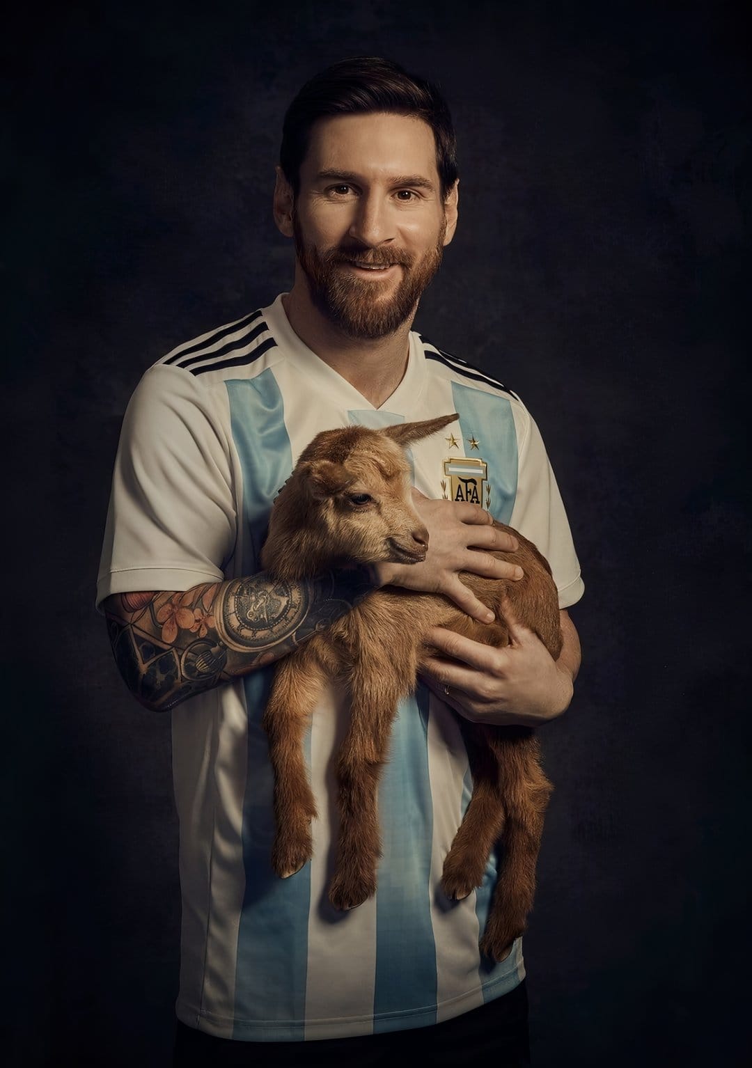 Lionel Messi cradles a brown baby goat while wearing a blue and white striped Argentina national football jersey.