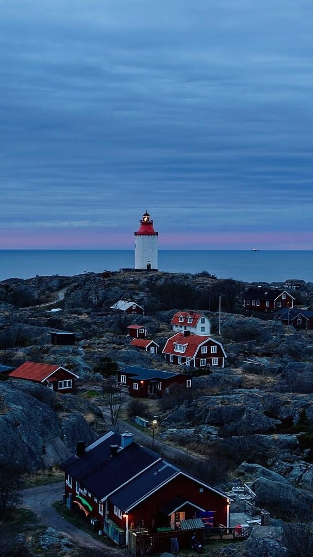 White lighthouse with a red lantern room overlooks small cottages and calm sea during a pink and blue twilight.