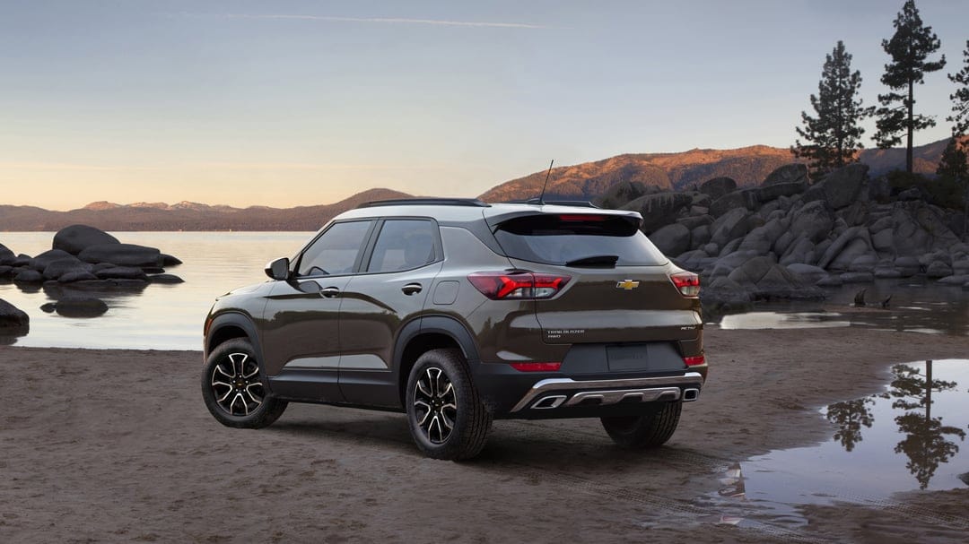 Metallic bronze SUV parked on a sandy beach beside a calm lake with mountains and pine trees at dusk.