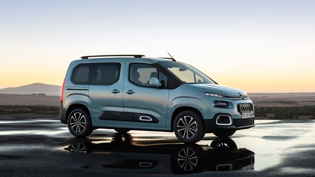 Teal Citroen Berlingo van parked on wet pavement reflecting a golden sunset sky over distant hills.