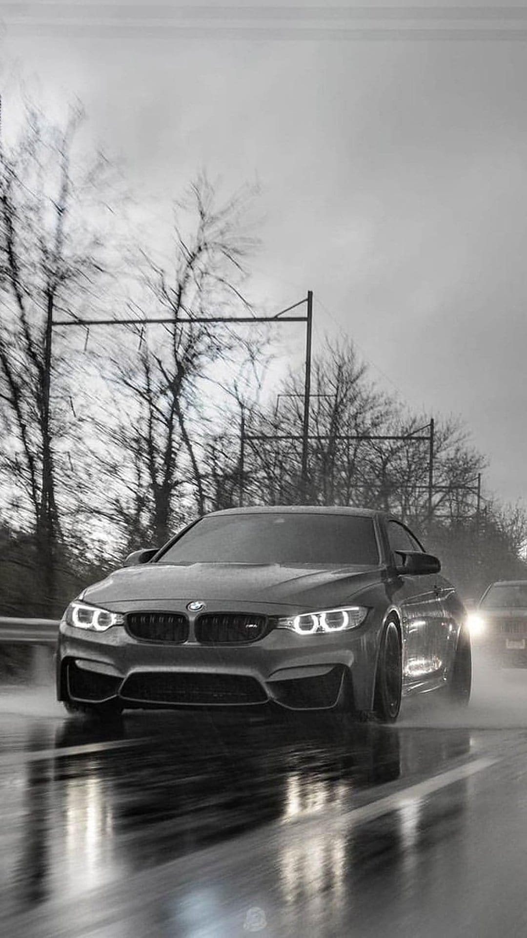 BMW M4 coupe driving on a wet road in the rain with glowing headlights and splashing water in black and white.
