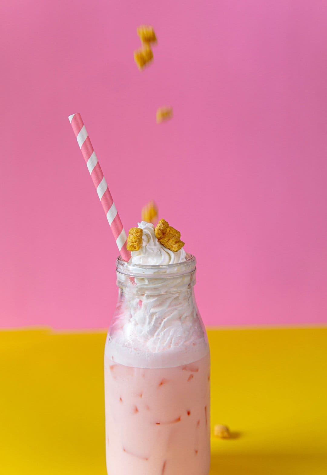 Strawberry milkshake in a glass bottle with whipped cream and cereal against a pink and yellow background.
