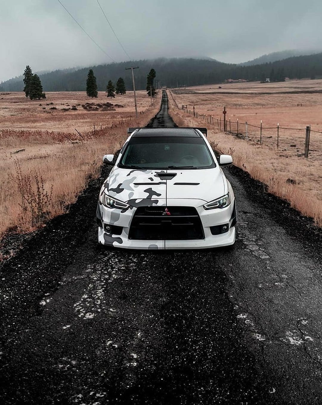 Mitsubishi Lancer Evolution with camo livery sits on a dirt road against misty hills and a gray sky.