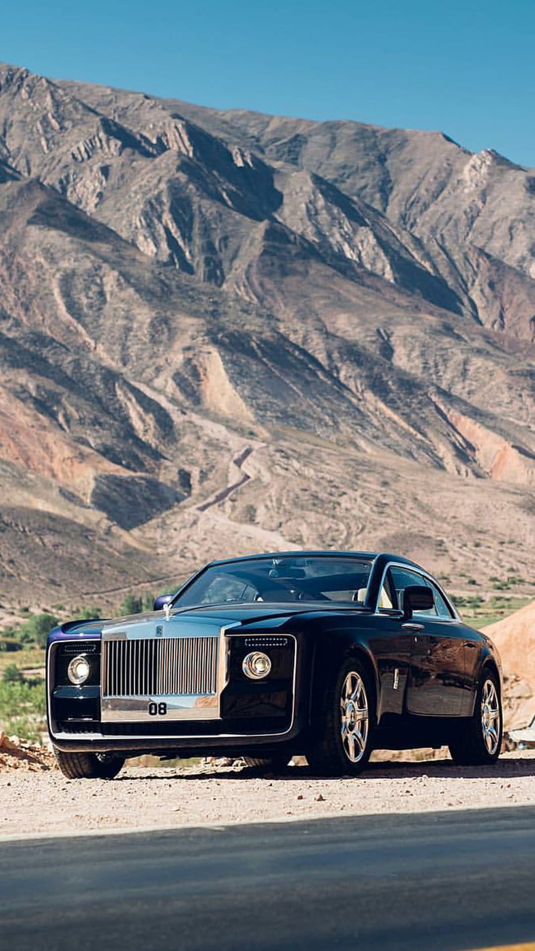 Black Rolls-Royce Phantom coupe parked on arid terrain with dramatic mountain peaks under a clear blue sky.