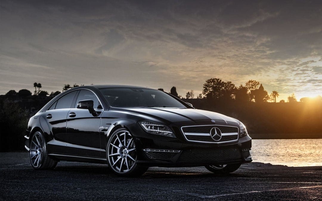 Black Mercedes-Benz CLS 63 AMG luxury sedan parked near a lake during a golden hour sunset with a cloudy sky.