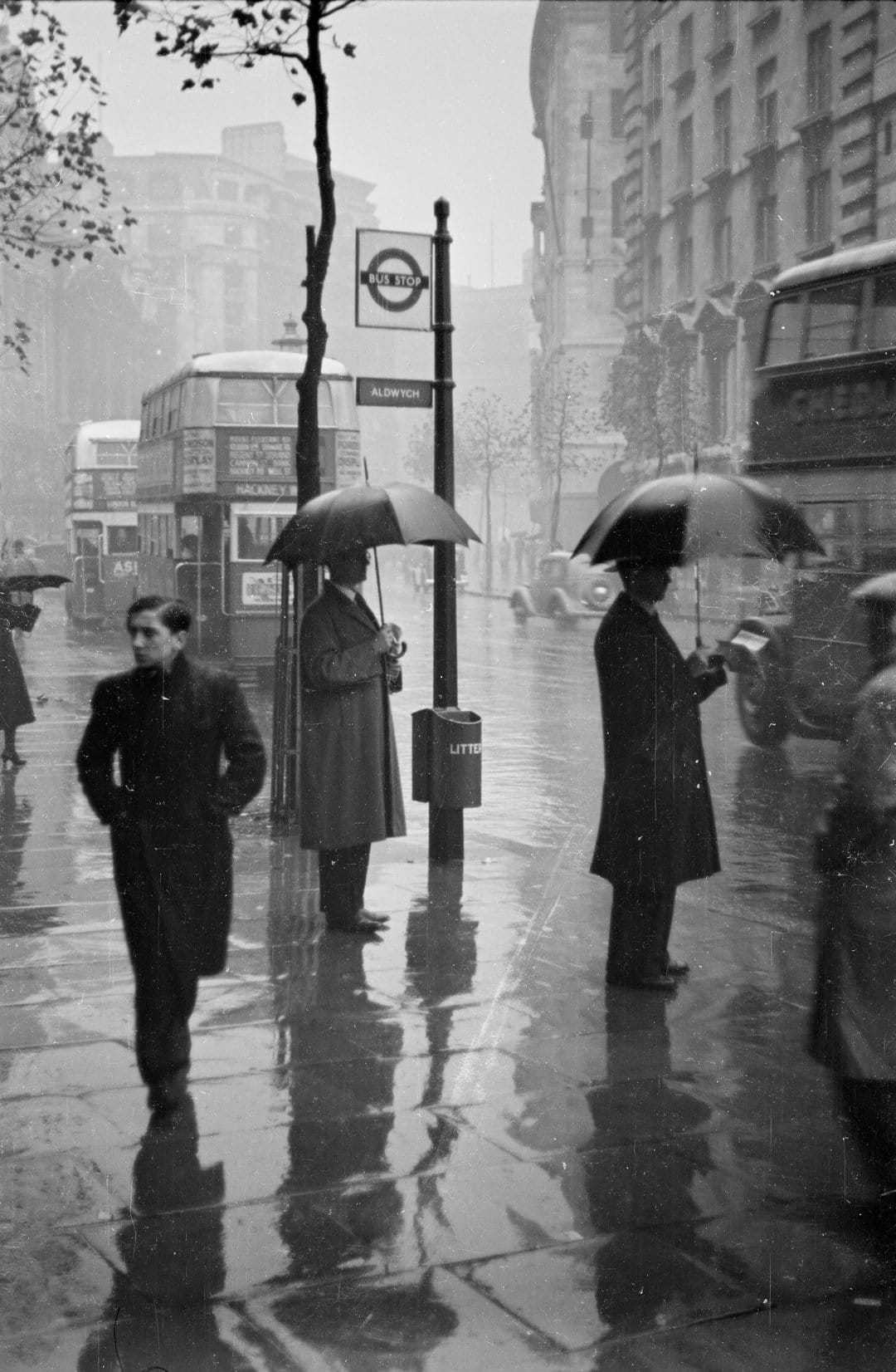 Pedestrians with umbrellas at a rainy London bus stop with reflections and a vintage Underground sign.
