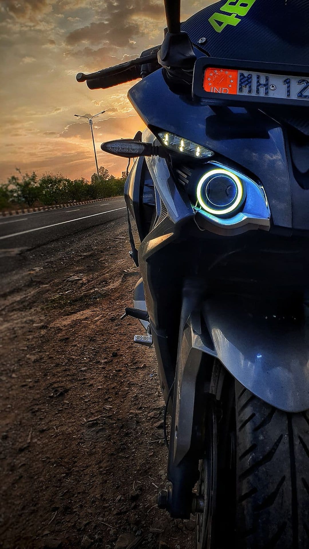 Dark blue motorbike front with a glowing halo LED headlight on a dusty roadside during a golden hour sunset.