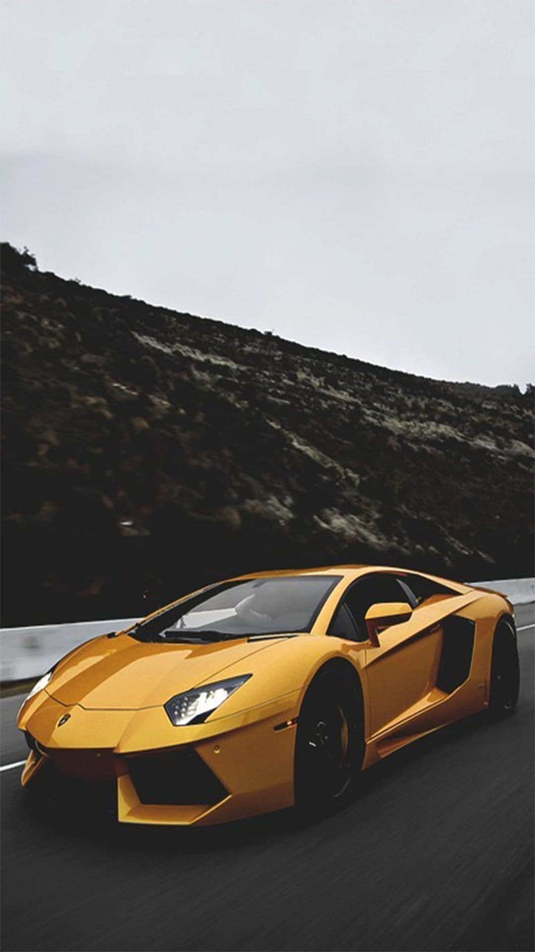 Golden Lamborghini Aventador racing on a mountain road under a cloudy sky with dark rocky peaks behind it.