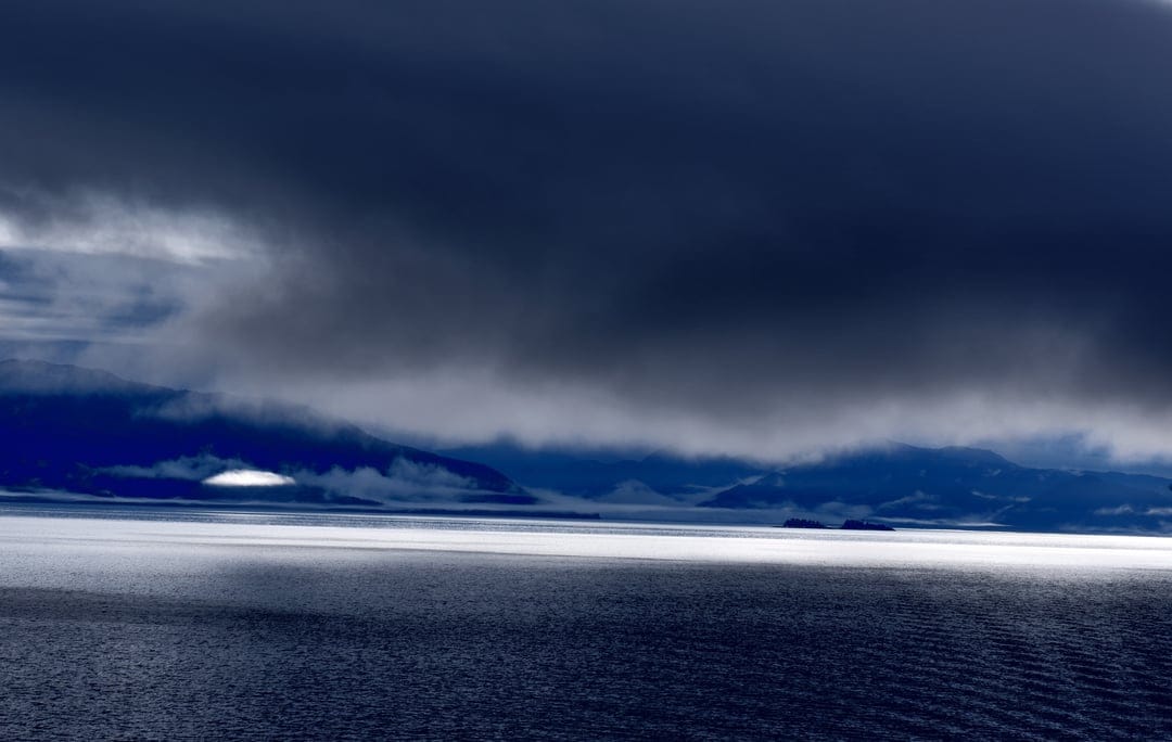 Heavy dark clouds hang above a rippling Arctic ocean with distant mountains and a thick layer of white sea mist.