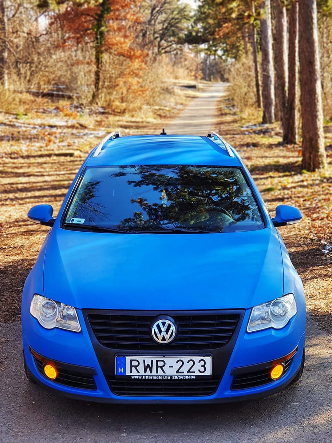 Blue Volkswagen Passat station wagon on a dirt road among orange autumn trees and seasonal forest foliage.