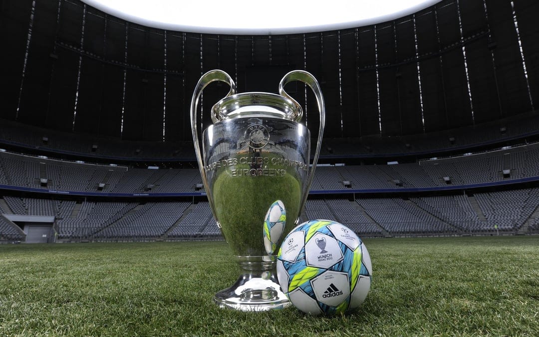 Silver UEFA Champions League trophy and star-patterned soccer ball on a green grass pitch under stadium lights.