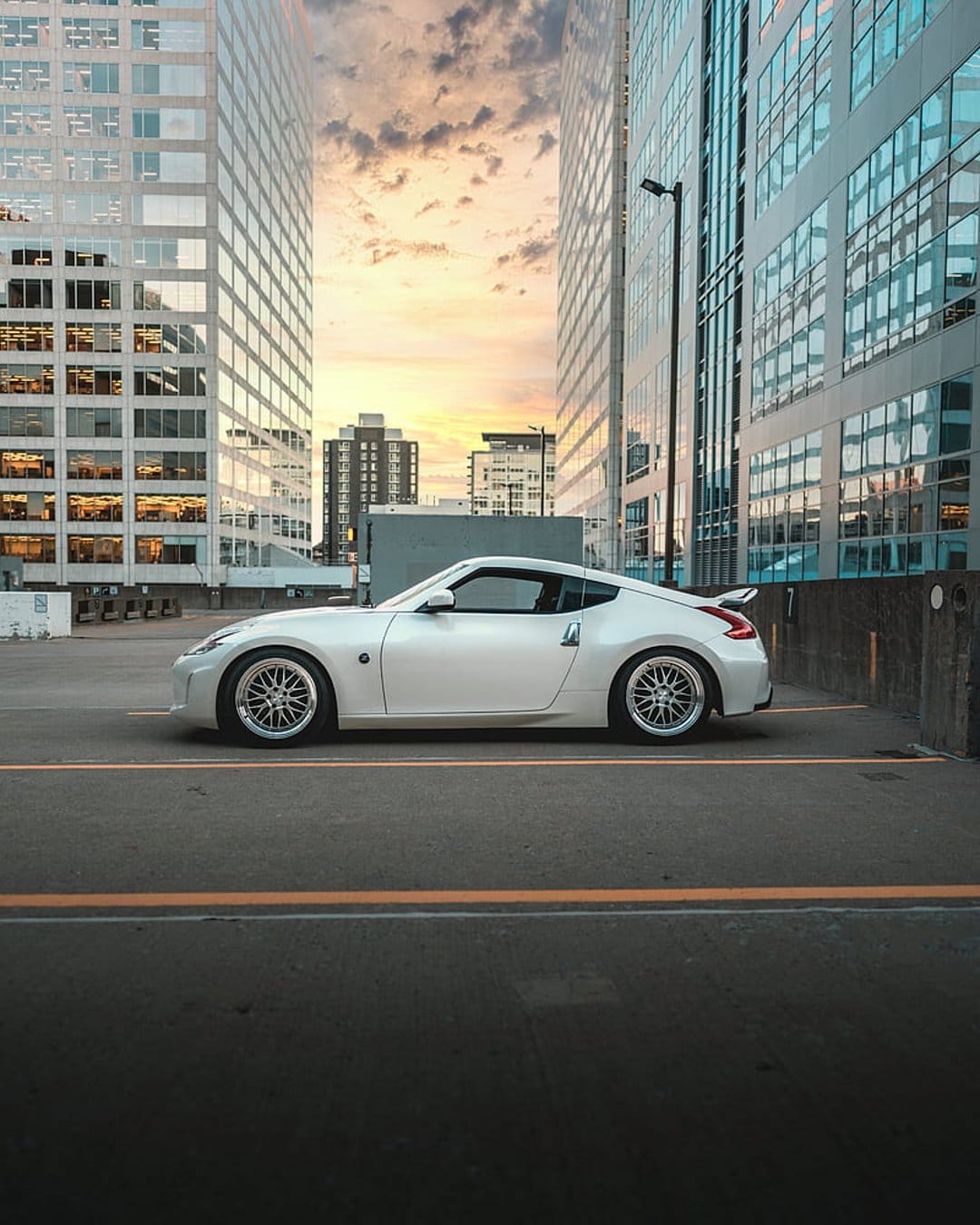 Nissan 370Z sports car parked between glass skyscrapers during a warm sunset with dramatic orange clouds.