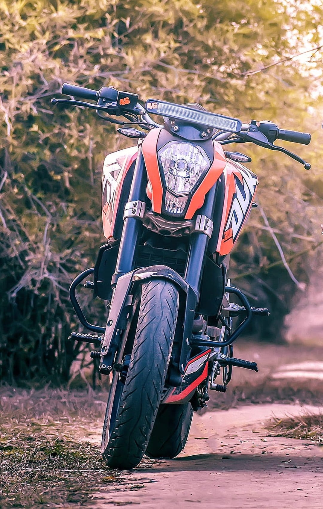 KTM Duke motorcycle with orange fairings and a thick front tire parked on a dirt path with green forest bokeh.