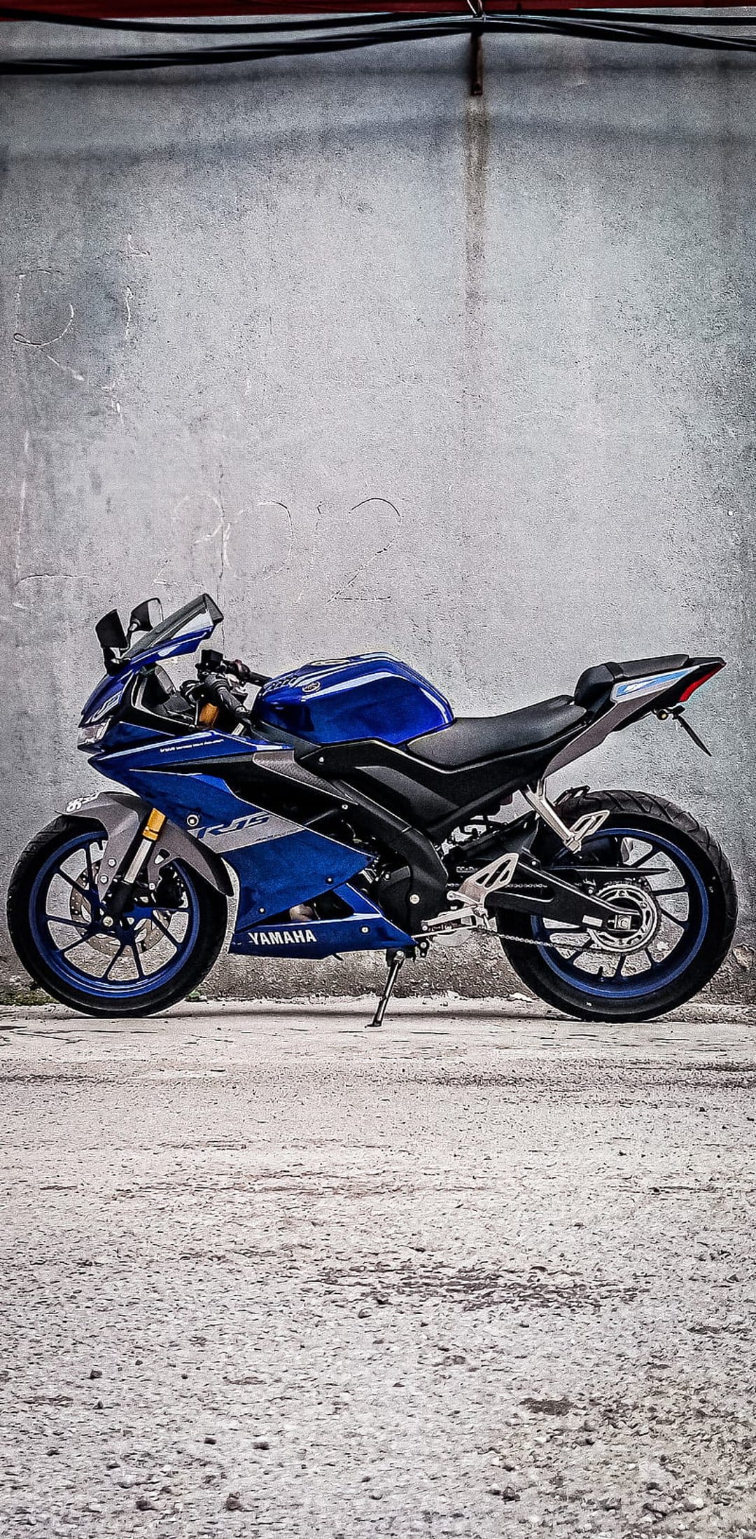 Blue Yamaha R15 motorcycle parked sideways against a textured gray concrete wall with overhead industrial pipes.