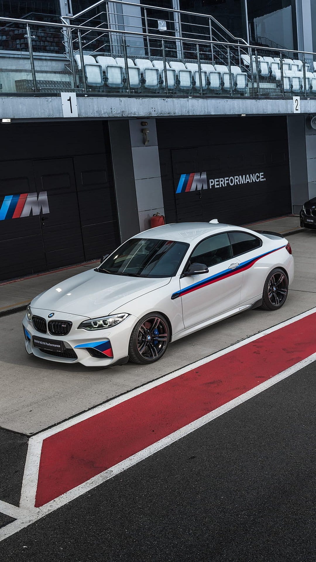White BMW M Performance car with M stripes parked on a race track pit lane near red and white asphalt markings.