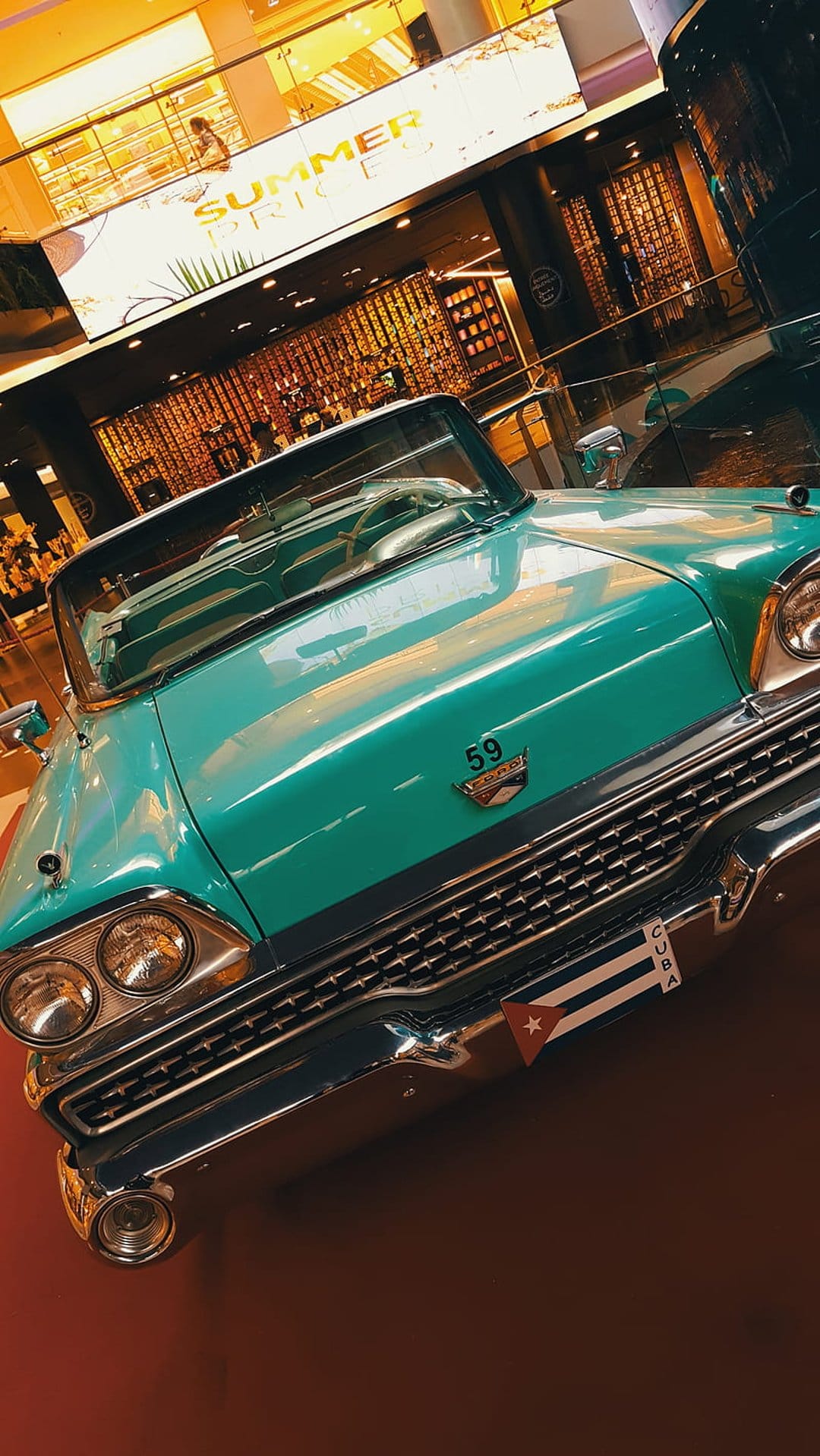 Teal 1950s convertible car with chrome grille and Cuba flag sticker on the bumper in a warm exhibition hall.