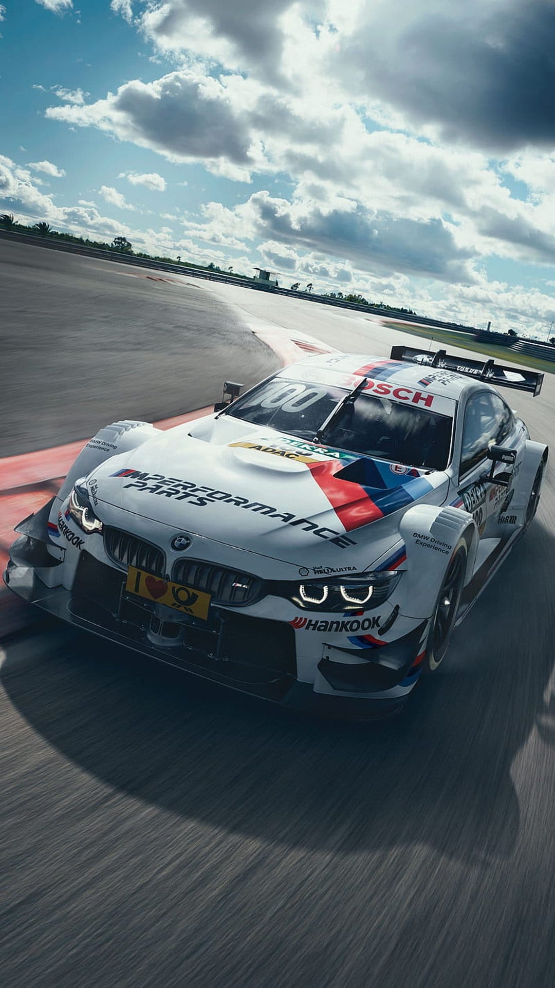 White BMW M Performance race car cornering a track under dramatic clouds with Bosch and ADAC sponsorship logos.
