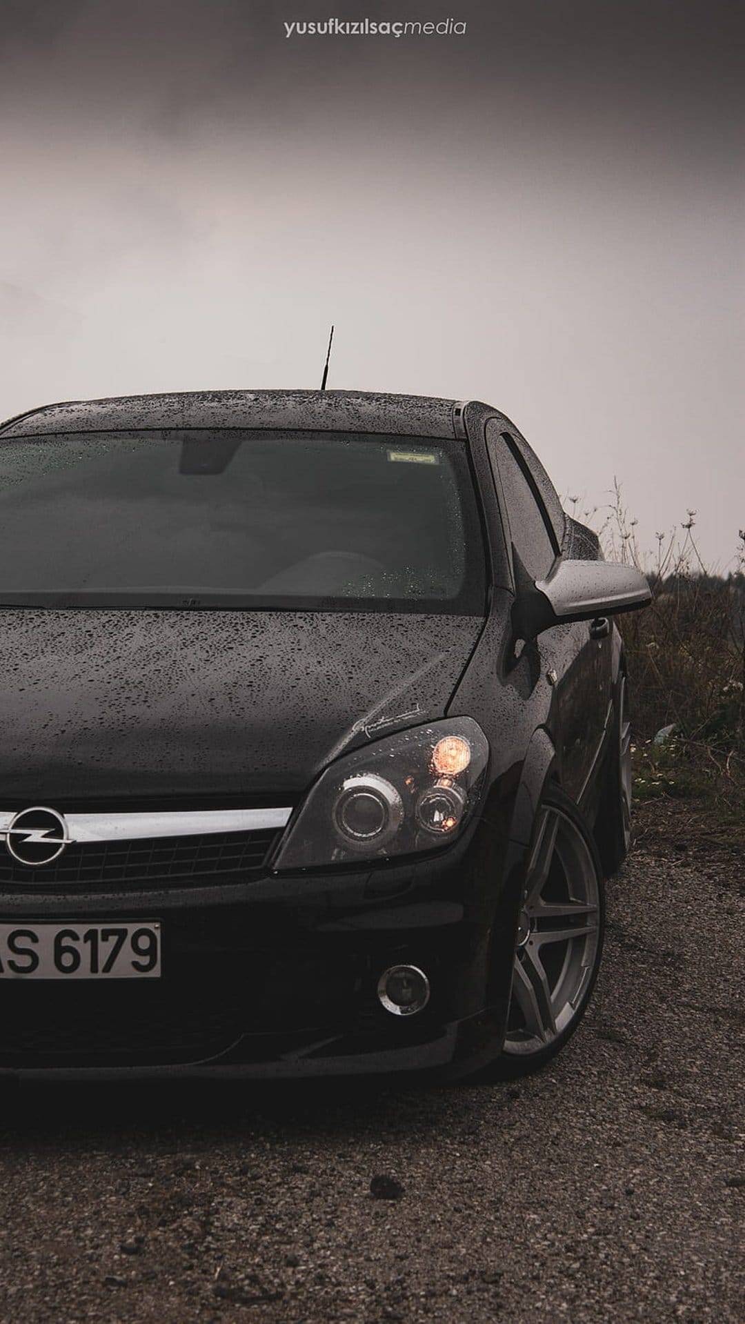 Black Opel Astra coupe parked on wet gravel with illuminated headlights under a cloudy, overcast sky