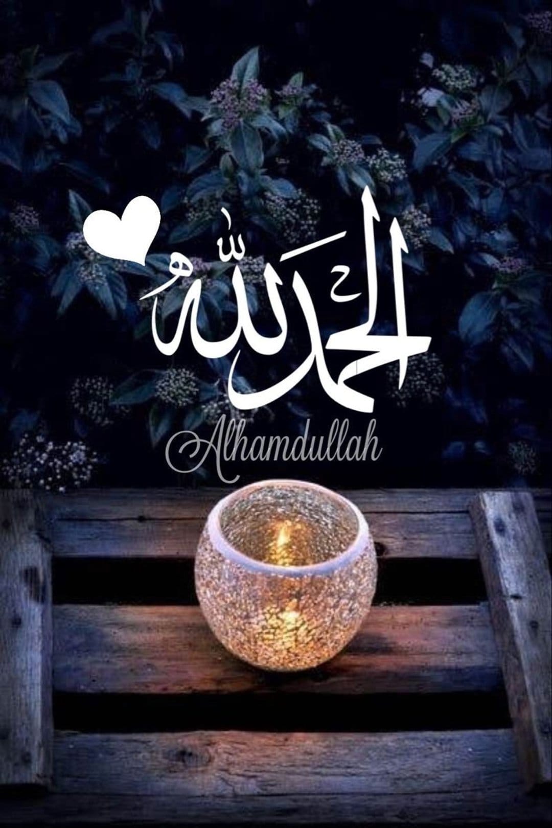 White Alhamdulillah calligraphy glows above a lit candle on a rustic wood surface with dark foliage behind.