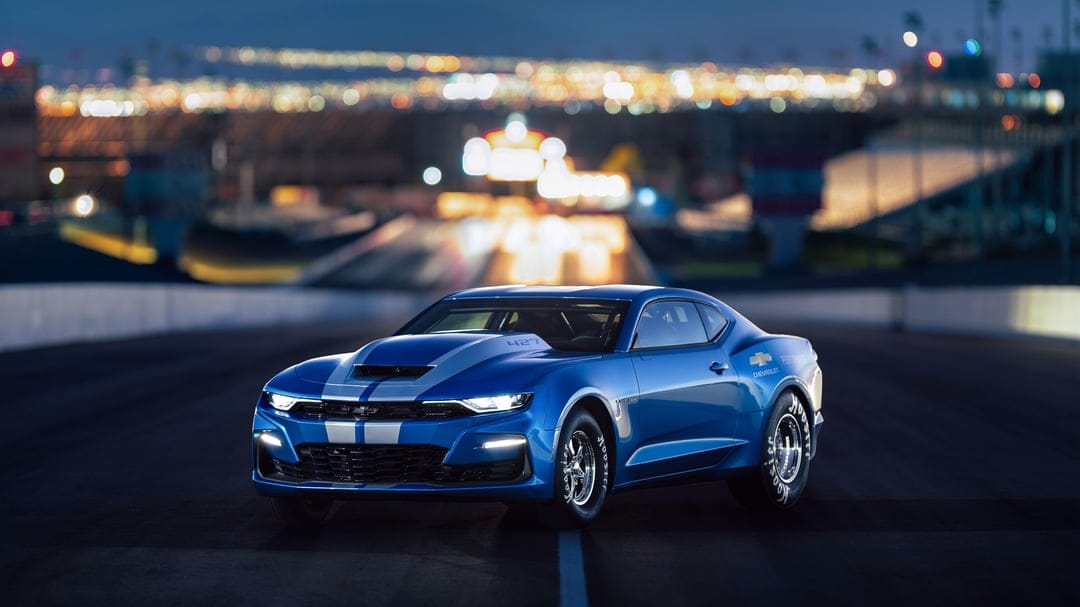 Blue Chevrolet COPO Camaro with silver stripes on a night track featuring soft bokeh city lights background.