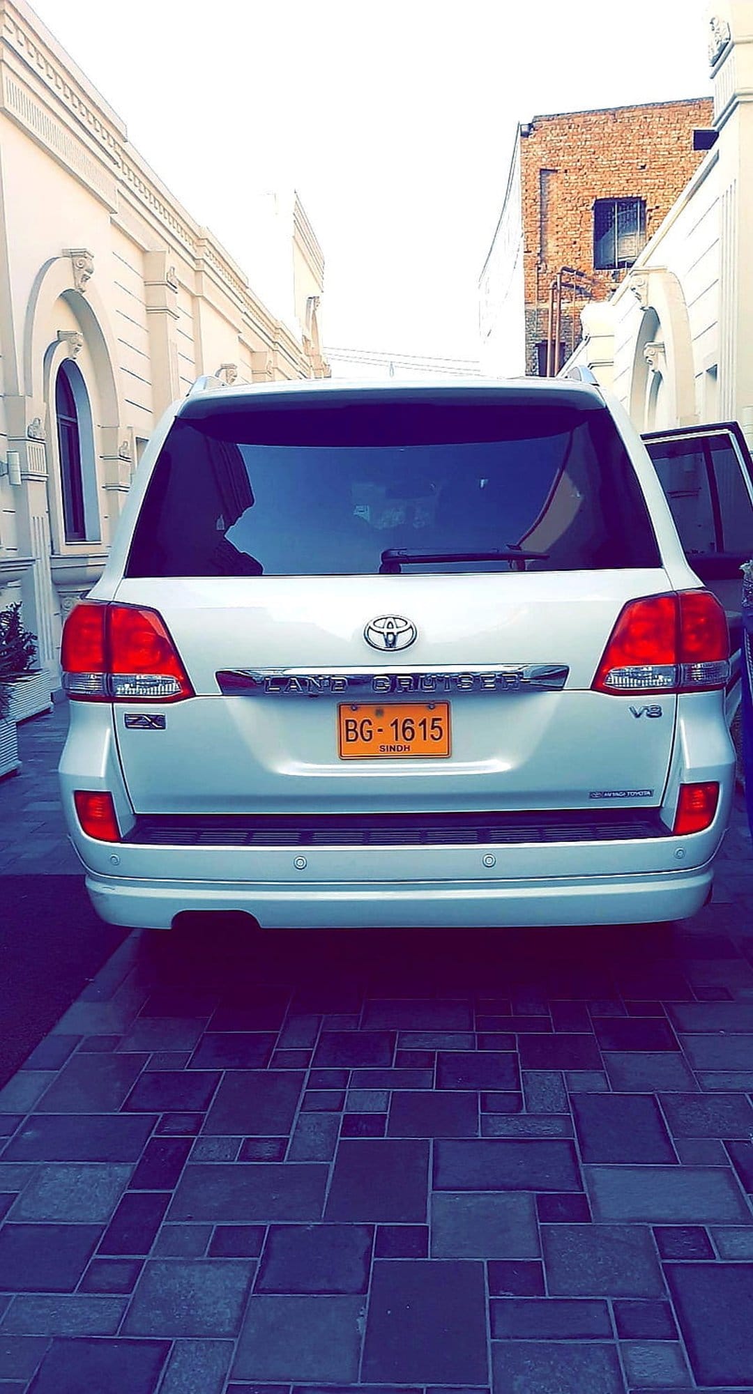 White Toyota Land Cruiser SUV parked on a tiled driveway between buildings with a yellow license plate visible.