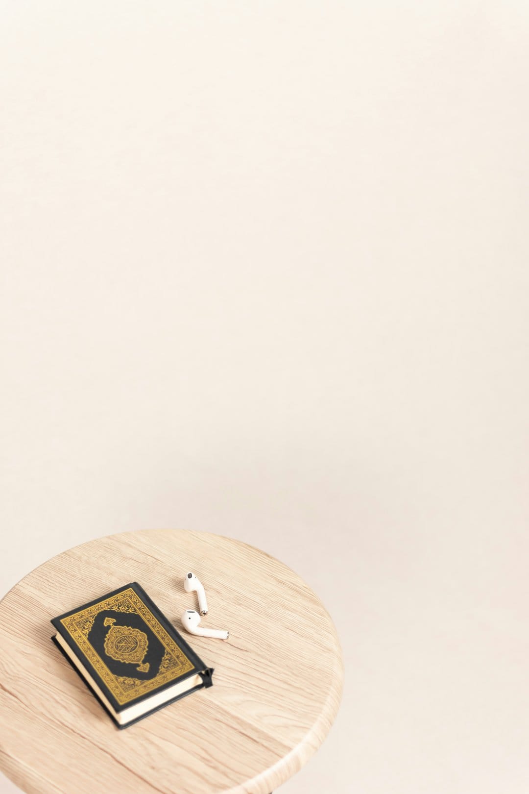 Open Quran with gold calligraphy beside white wireless earbuds on a light wood table against a cream background.