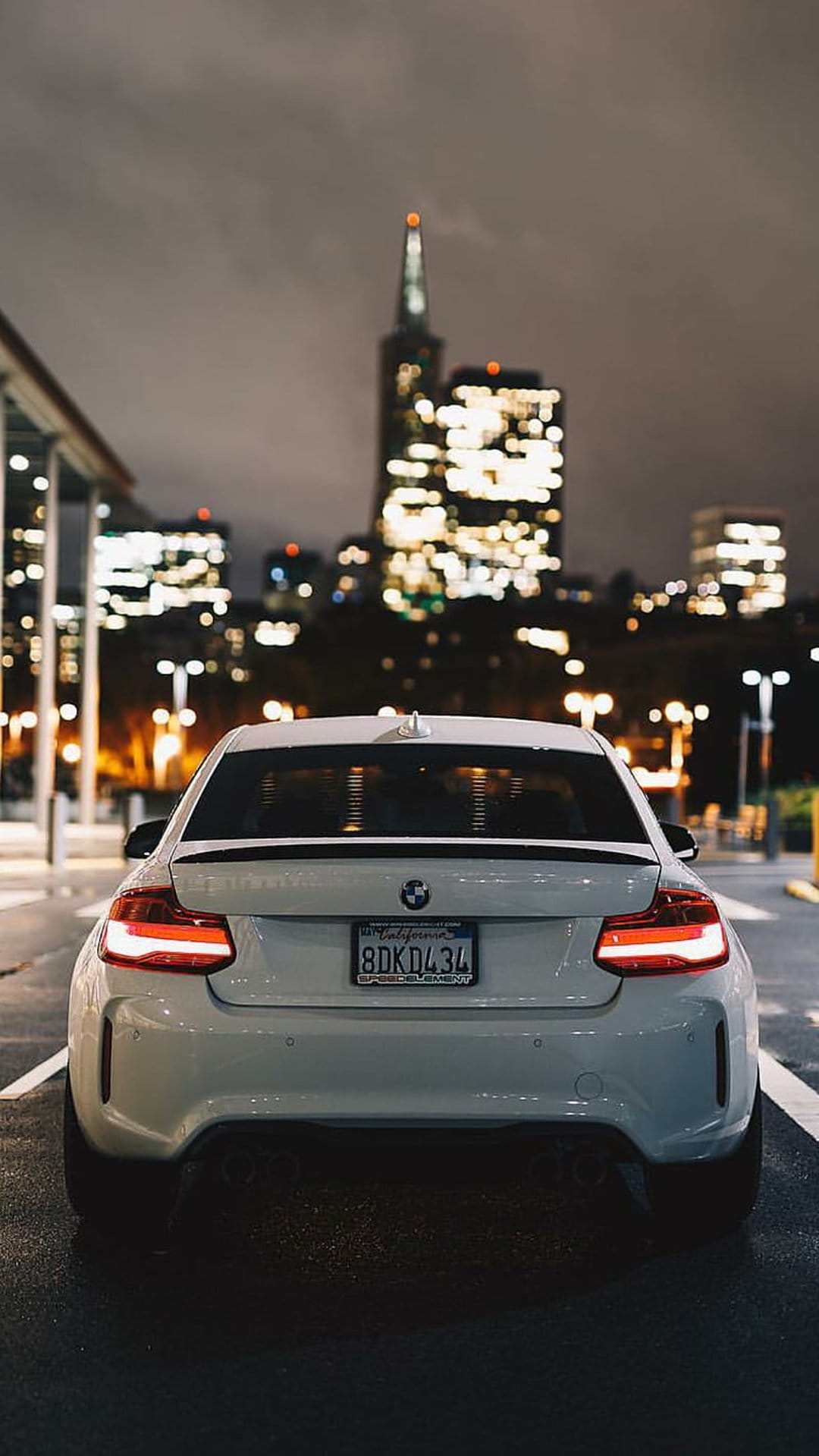 White BMW coupe rear view on wet asphalt with red taillights glowing against a distant city skyline at night.
