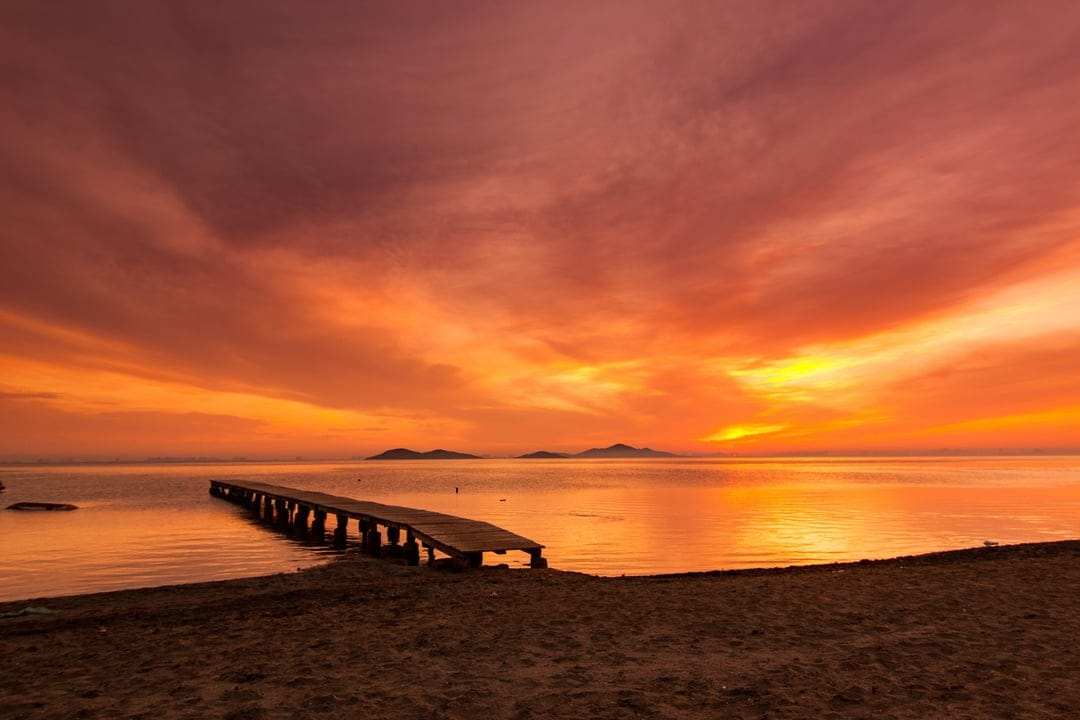 Weathered wooden pier leading into a calm sea under a vibrant orange and red sunset sky with distant islands.