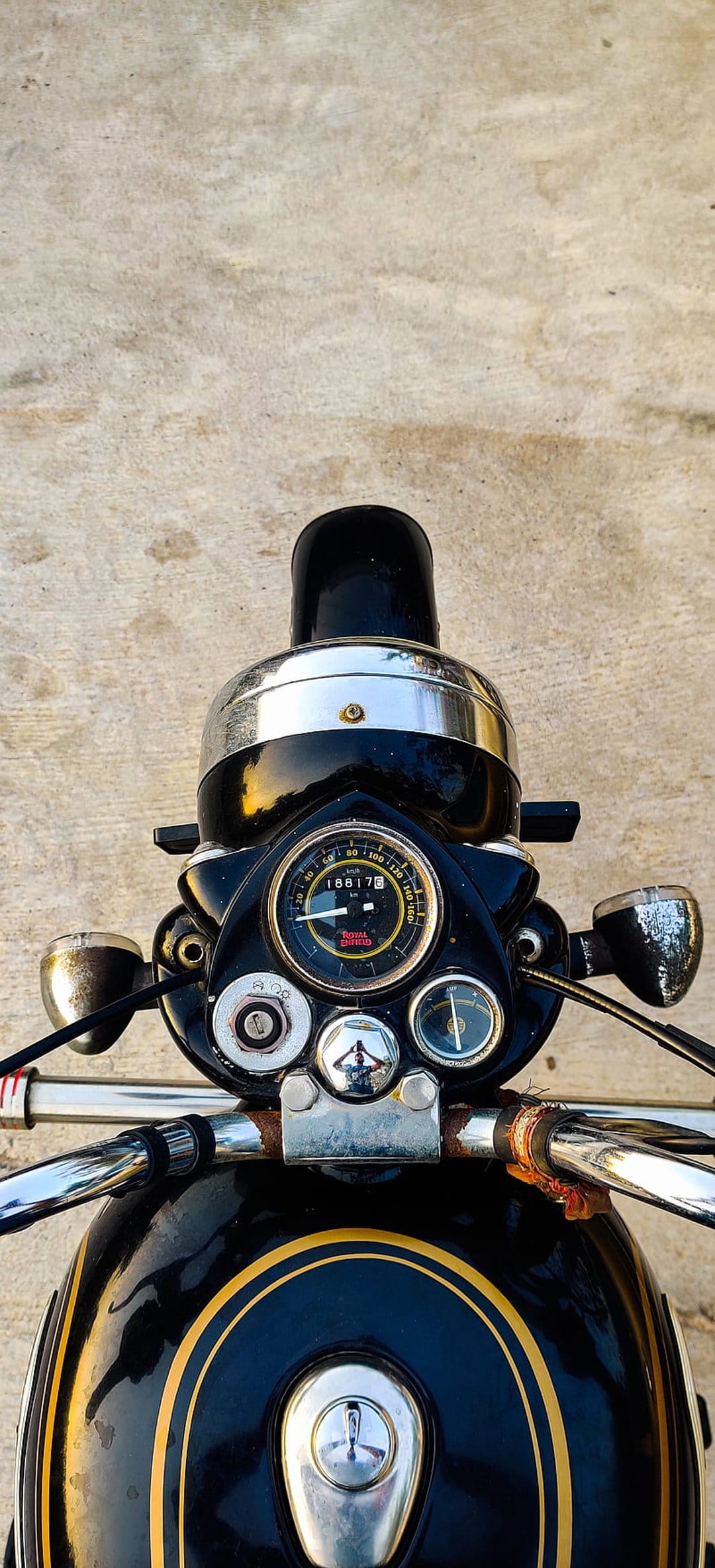 Royal Enfield speedometer and chrome handlebars on a black fuel tank with gold pinstriping in a close-up shot.