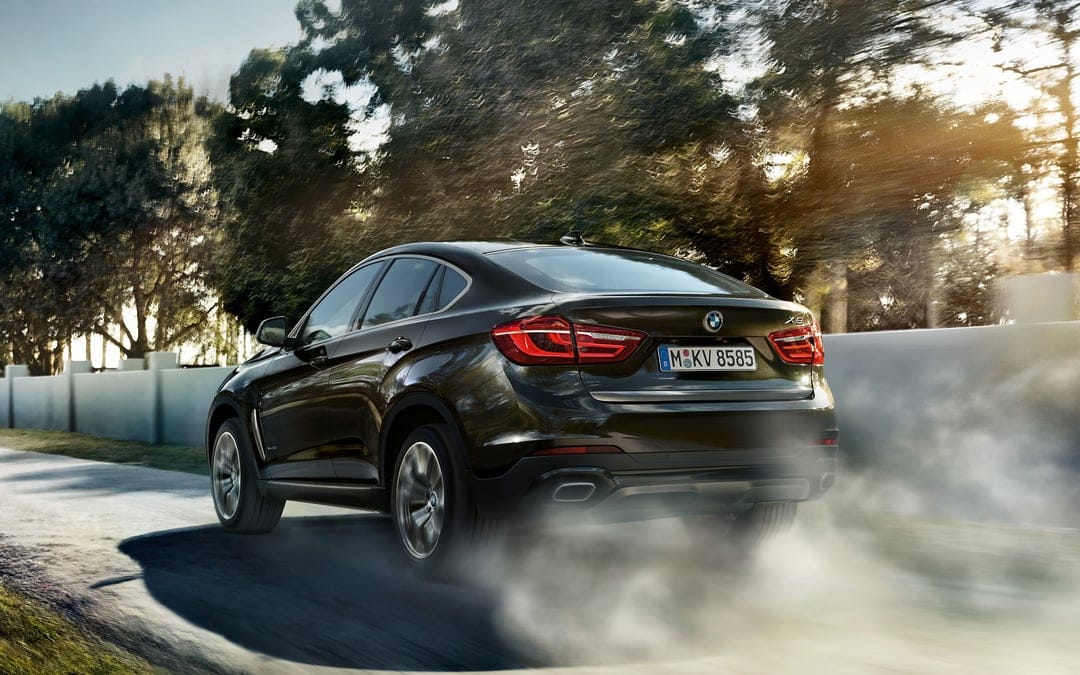 Black BMW X6 luxury SUV driving fast on a road, kicking up white smoke under golden sunlight through trees.
