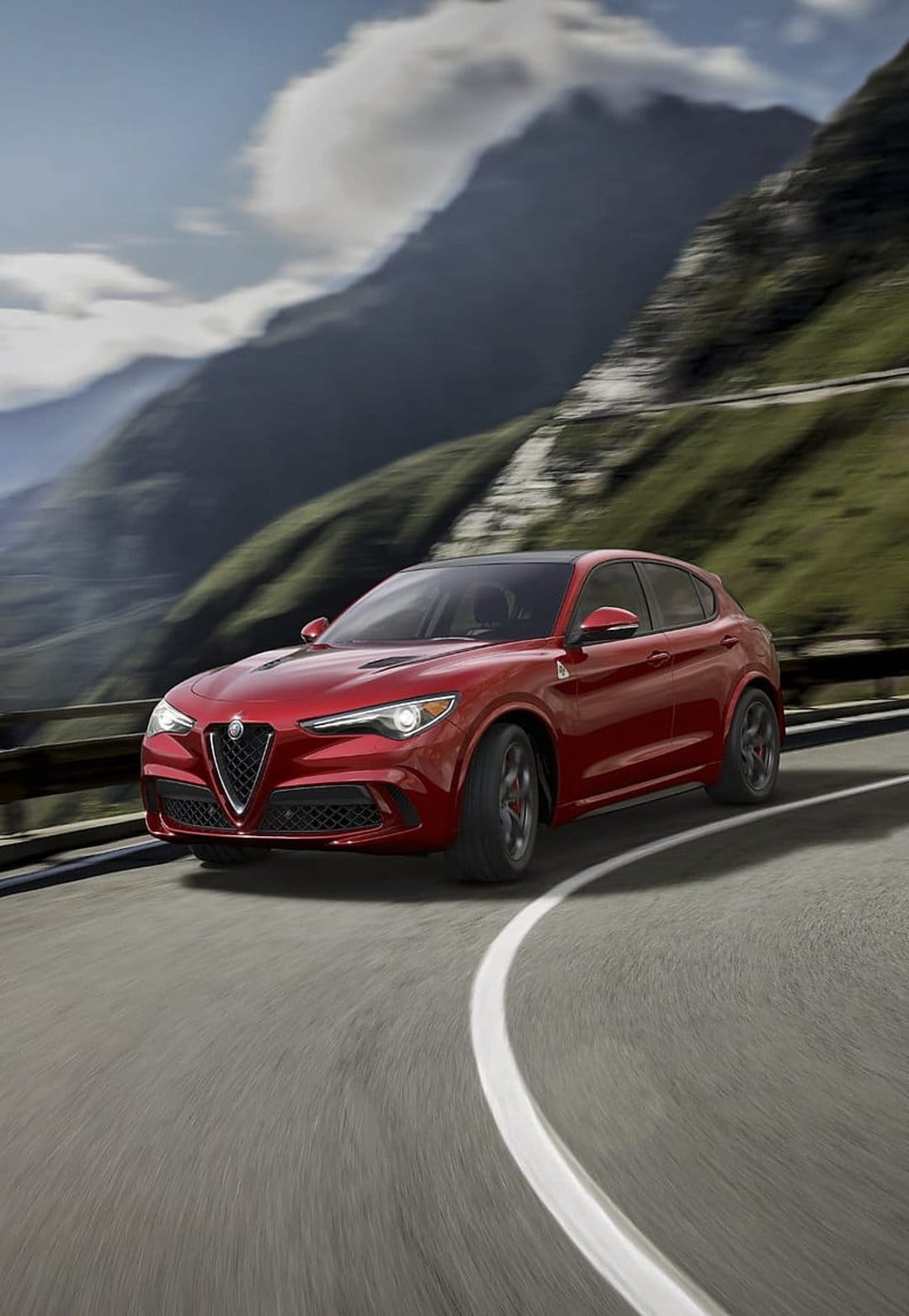 Red Alfa Romeo Stelvio SUV driving fast on a curved mountain road with blurred green hills and bright blue sky.