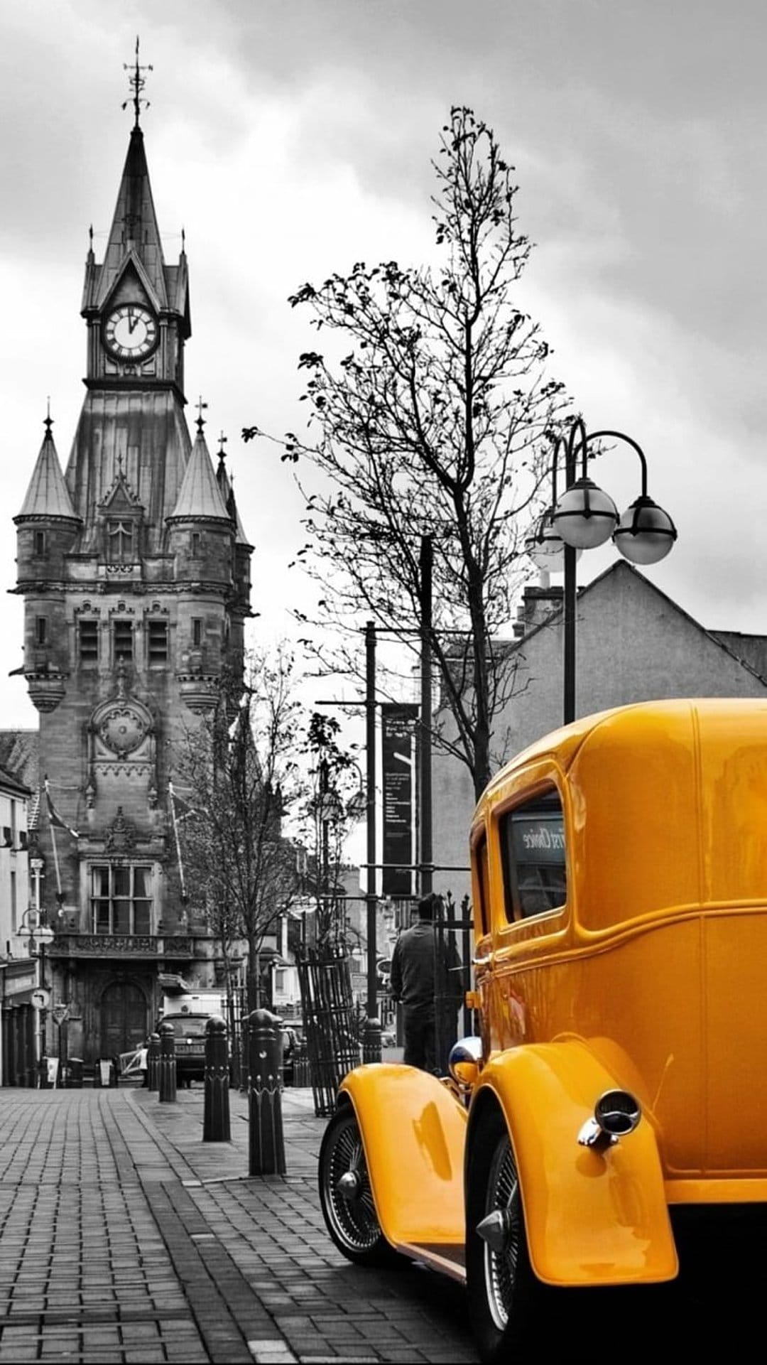 Yellow vintage car parked on a monochrome cobblestone street in front of a tall historic stone clock tower.