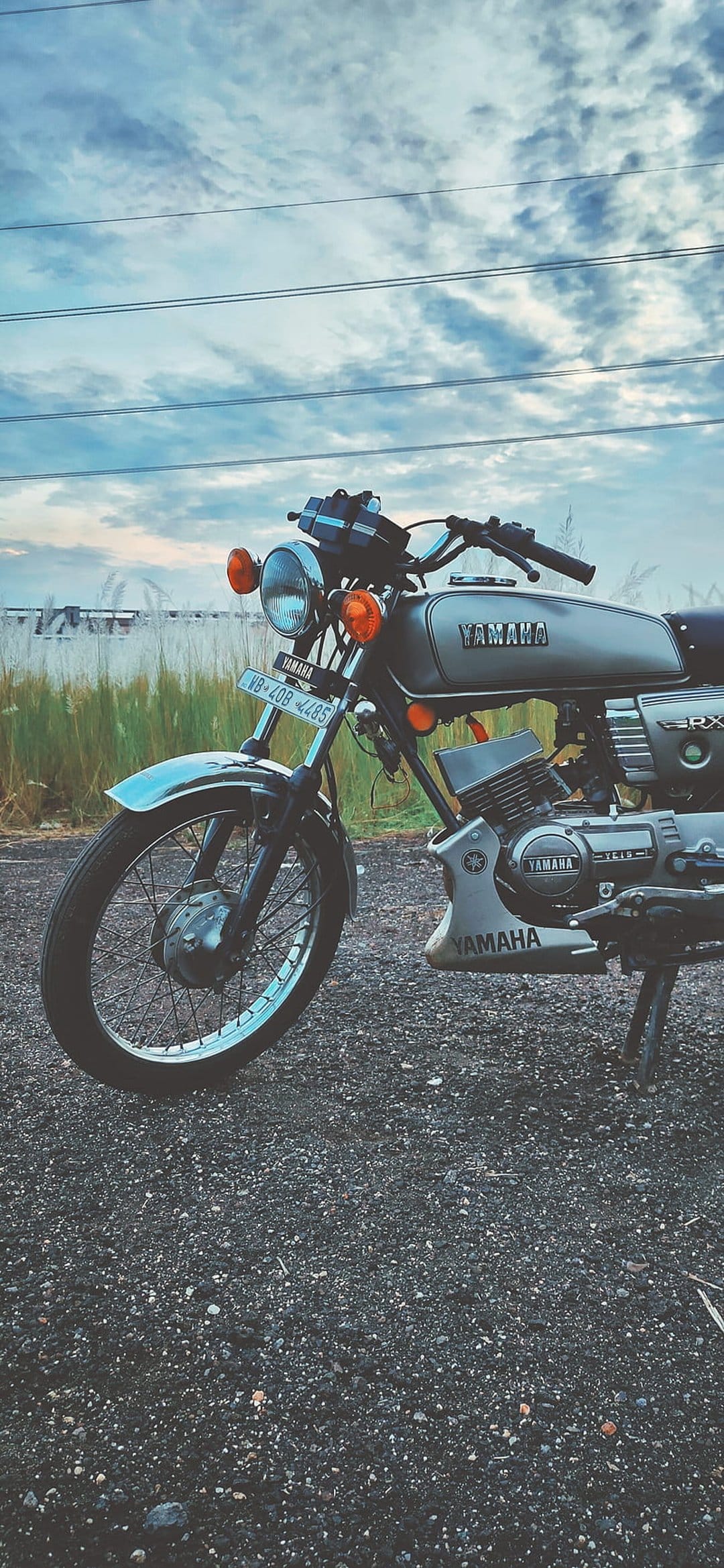 Yamaha RX100 silver motorcycle parked on a gravel path against tall grass and a cloudy sky in a vertical frame.