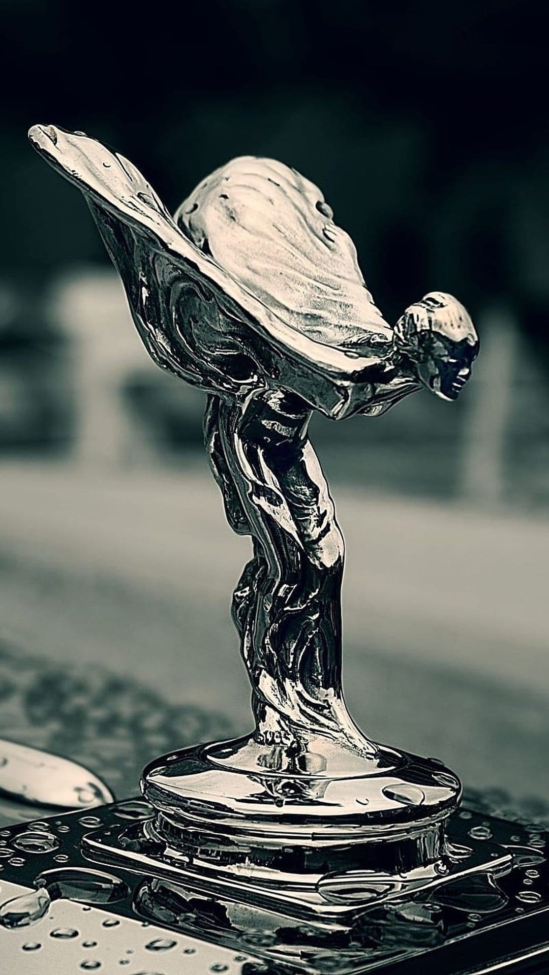 Spirit of Ecstasy sculpture with water droplets on polished chrome captured in a monochrome vertical portrait.