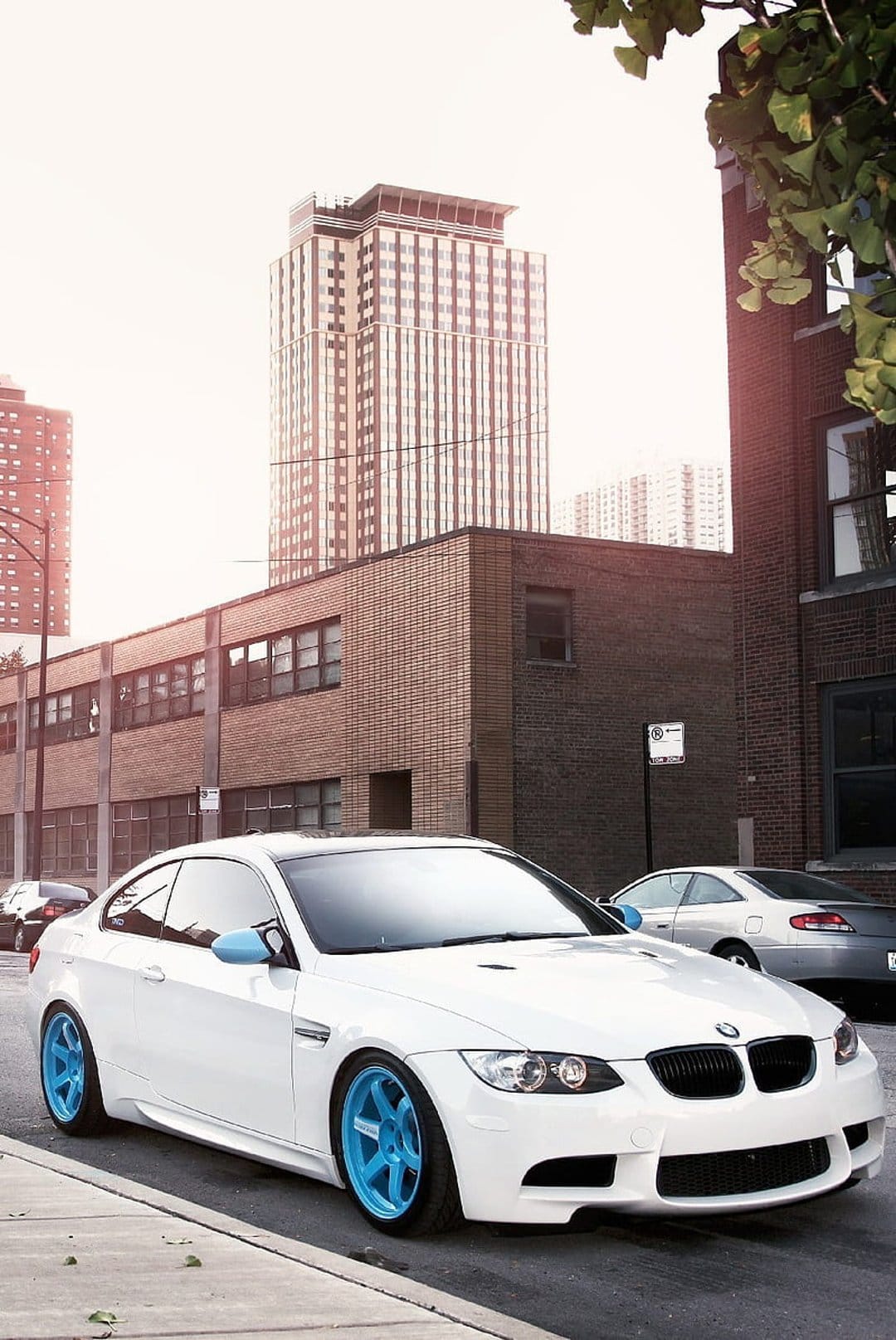 White BMW coupe with bright blue rims parked on a city street between brick buildings and tall skyscrapers.