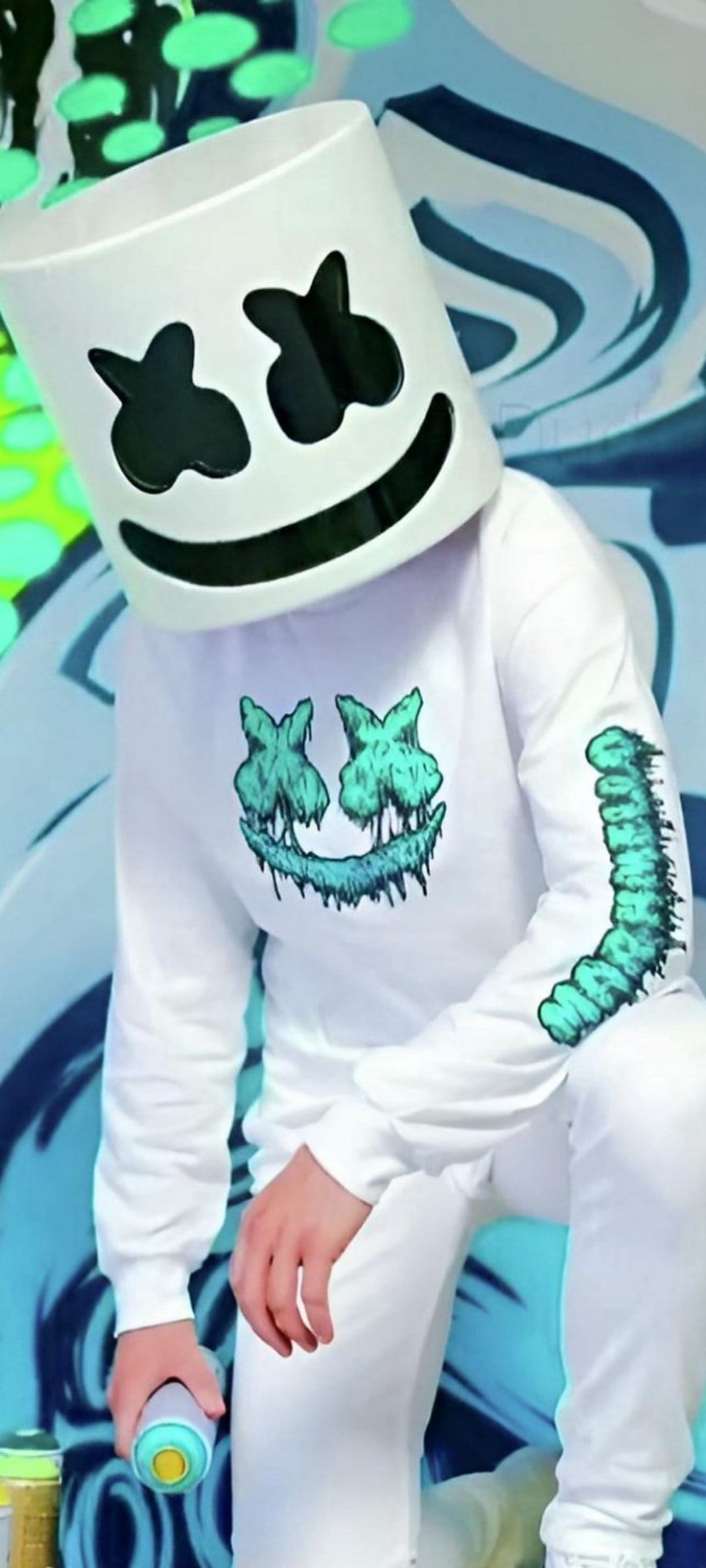 Marshmello in a white helmet and teal-stained hoodie holds a spray can before a blue and green abstract mural.